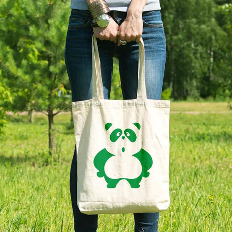 Person outdoors holding a tote bag with a green panda graphic, standing in grass with trees in background, wearing jeans and bracelet.