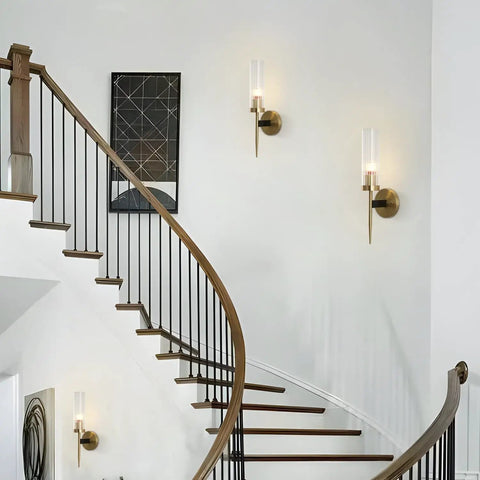 Mid Century Modern Gold Wall Light With Cylinder Glass Shade Brushed Brass Finish Long Wall Sconce For Vintage Bathroom And Hallway Mount Lamp With Lit Glow And Classic Vibe in curved staircase setting.