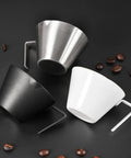 Three espresso cups, including a Two Piece Stainless Steel Espresso Cups With Handles And A Pro Pour Double Spout Measuring Cup With Dual Scale V Shaped Mouth For Drip Free Espresso Shots 3.4 Oz 100 Ml Silver, black and white, on a dark surface with scattered coffee beans.