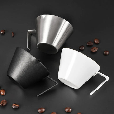 Three espresso cups, including a Two Piece Stainless Steel Espresso Cups With Handles And A Pro Pour Double Spout Measuring Cup With Dual Scale V Shaped Mouth For Drip Free Espresso Shots 3.4 Oz 100 Ml Silver, black and white, on a dark surface with scattered coffee beans.