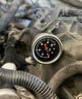 Inline fuel pressure gauge with round metal bezel and black dial, 0-100 psi, red needle mounted in engine bay.