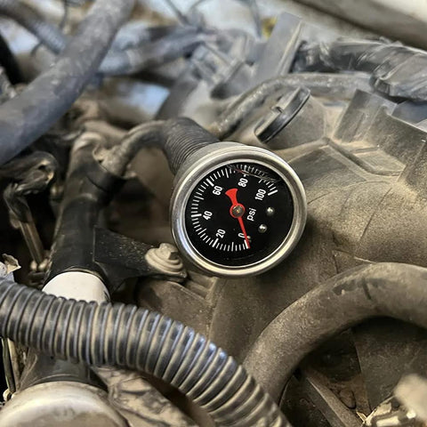 Inline fuel pressure gauge with round metal bezel and black dial, 0-100 psi, red needle mounted in engine bay.