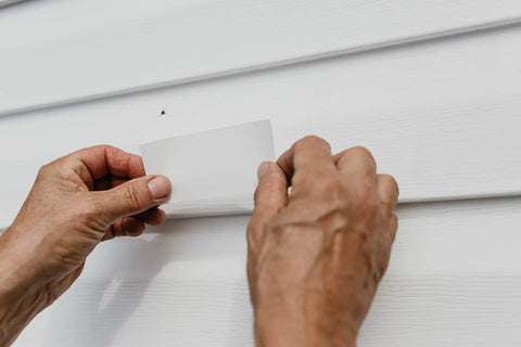 Hands applying patch from Vinyl And Stucco Siding Repair Kit With White Self Adhesive Patches That Cut To Fit Paintable To Match Cover Cracks Holes And Blemishes Weather Resistant Indoor Outdoor Use Includes 2 Patches on white siding.