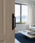 Close-up of black barn door handle on wooden door with modern living room background, highlighting 12 Pack Black Barn Door Handle Cabinet Pulls Aluminum Alloy 5.8 Inch Long With Screws For Kitchen Cabinets Cupboards Wardrobes Closets Sheds Garages And Sliding Barn Doors Perfect For Upgrading Drawer Pulls And Cabinet Hardware.