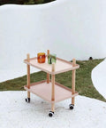 Pink two-tier cart with wooden frame and caster wheels holding two colorful cups on a minimalist white and green background.