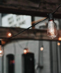 25 Pack Edison Style Replacement Light Bulbs 7-Watt E12 Candelabra Base ST40 Clear Glass Warm White 2300K Ideal For Outdoor Patio String Lights Waterproof And Rust Resistant hanging in a rustic patio setting.