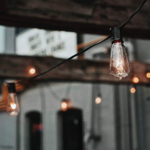 25 Pack Edison Style Replacement Light Bulbs 7-Watt E12 Candelabra Base ST40 Clear Glass Warm White 2300K Ideal For Outdoor Patio String Lights Waterproof And Rust Resistant hanging in a rustic patio setting.