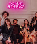 Group celebrating beneath This Must Be The Place Neon Sign Pink Wall Decor For Bedroom And Party Vibes, 23 by 9 inches.