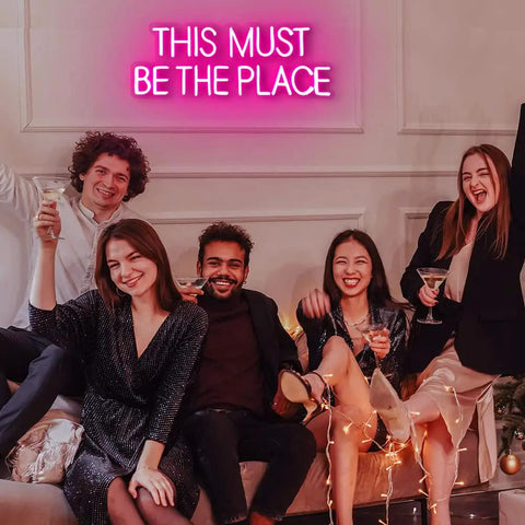 Group celebrating beneath This Must Be The Place Neon Sign Pink Wall Decor For Bedroom And Party Vibes, 23 by 9 inches.