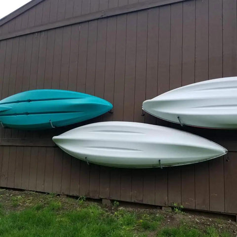 Beefy Wall Mount Kayak Storage Rack With Lashing Straps, Heavy Duty Garage Hanger For Kayaks, SUPs, Paddle Boards And Surfboards, 100 Lb Capacity, EVA Foam Protected Arms, Easy Access Hooks, Black And Red installed holding three kayaks on an outdoor wall.
