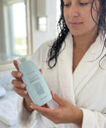 Woman in a robe holding Quick Fix Leave In Hair Treatment bottle in a bright bathroom setting.