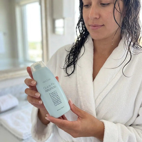 Woman in a robe holding Quick Fix Leave In Hair Treatment bottle in a bright bathroom setting.