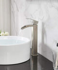 Modern Vessel Sink Faucet In Brushed Nickel Tall Waterfall Style Bathroom Mixer Single Handle With Pop Up Drain And Three Eighths Inch Hoses Clean Minimalist Look For Contemporary Bathrooms next to white sink on black countertop.