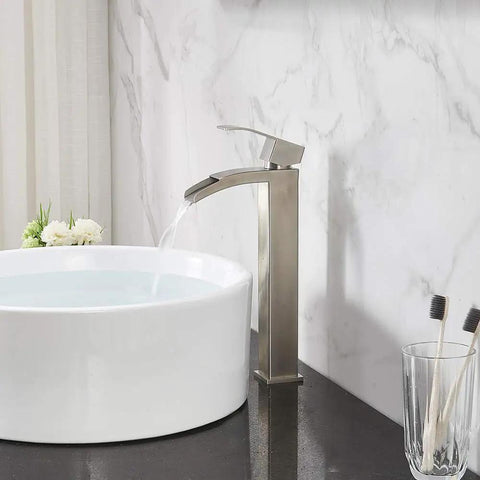 Modern Vessel Sink Faucet In Brushed Nickel Tall Waterfall Style Bathroom Mixer Single Handle With Pop Up Drain And Three Eighths Inch Hoses Clean Minimalist Look For Contemporary Bathrooms next to white sink on black countertop.