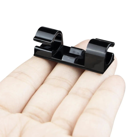 Hand holding a black clip from 50 Piece Self Adhesive Cable Management Clips Set For TV PC Laptop And Ethernet Cables No Mess Sticky Wire Clips Cord Holder Easy Tidy Organization For Desktop Home Office.