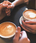 Close-up of two people holding cappuccino cups with latte art at a cafe table.