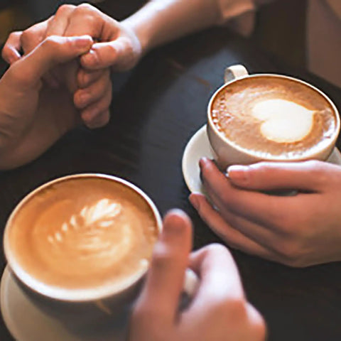Close-up of two people holding cappuccino cups with latte art at a cafe table.