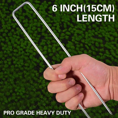 Hand holding heavy duty 6 inch garden landscape staple, U shaped, 11 gauge galvanized pin, pro grade visible on grass surface.