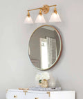 Three Light Gold Vanity Light Fixtures With White Frosted Glass Shades Over Mirror In Brushed Antique Gold Finish