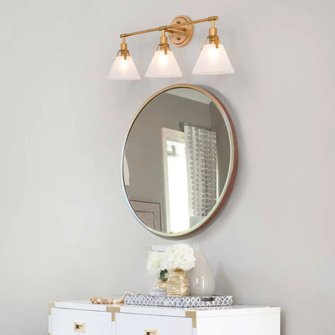 Three Light Gold Vanity Light Fixtures With White Frosted Glass Shades Over Mirror In Brushed Antique Gold Finish