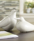 Two white ceramic bird figurines on a tabletop with a blurred background of kitchen decor and folded napkin in foreground.