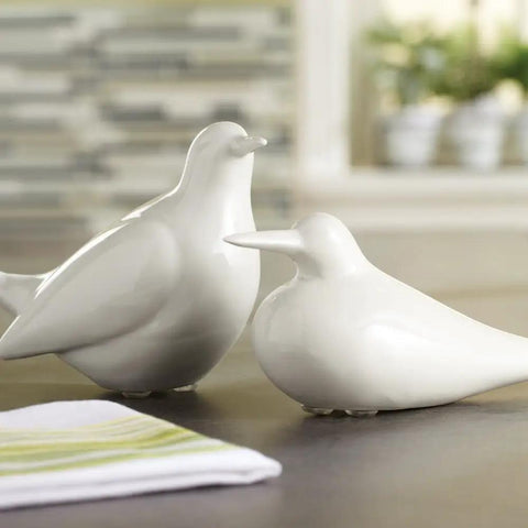 Two white ceramic bird figurines on a tabletop with a blurred background of kitchen decor and folded napkin in foreground.