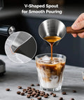 Stainless Steel Espresso Measuring Cup With Wide Mouth And V Shaped Pour Spout, Easy Grip Handle, Built In Scale For Latte Cappuccino Americano Macchiato, Pro Grade For Home Baristas Black pouring espresso into iced coffee.