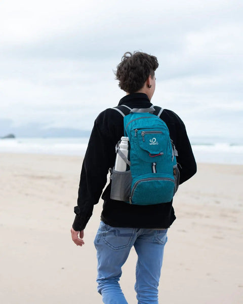 Person walking on the beach with Ultra Lightweight 20L Packable Daypack For Travel, Hiking And Camping, Foldable And Compact, teal color.