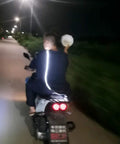 Person riding a motorcycle at night on a road, holding an object, wearing a reflective jacket, with streetlights visible.
