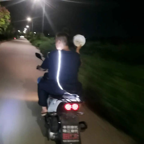 Person riding a motorcycle at night on a road, holding an object, wearing a reflective jacket, with streetlights visible.