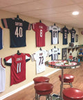 Wall with various jerseys displayed using Jersey Display Hanger For Wall Adjustable Pivot Joint That Shapes To Fit Any Sport