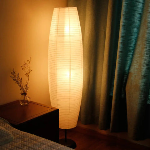 Foldable Rice Paper Floor Lamp Shade In Minimalist Modern Style next to a bed and nightstand in a softly lit bedroom, 43.3 cm tall.