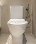 Front view of a toilet in a modern bathroom setting with beige tiles and a handheld bidet sprayer visible on the side.