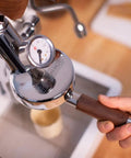 Barista using Bottomless 58mm Portafilter for E61 Grouphead Espresso Machines, Zebra Wood handle visible against stainless steel body.