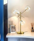 Modern Gold Table Lamp With 3 Light Glass Globe Bulbs, Antique Brass Finish on a desk beside a bed, showcasing mid-century design.