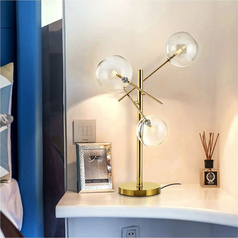 Modern Gold Table Lamp With 3 Light Glass Globe Bulbs, Antique Brass Finish on a desk beside a bed, showcasing mid-century design.
