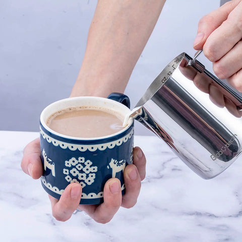 Pouring milk from Stainless Steel Milk Frothing Pitcher 12 Oz 350ml For Latte Art And Creamy Foam into a decorative mug.