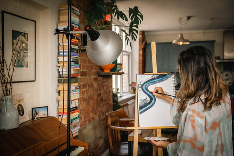 Artist uses lightweight studio lamp with foldable tripod and adjustable aluminum shade for painting on canvas indoors.