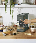 Rock Solid Compact Espresso Machine In Fresh Thyme Green with a 20 bar pump, on a kitchen counter, surrounded by coffee and snacks.