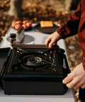 10,000 BTU Dual Fuel Camp Stove for Butane and Propane Portable Outdoor Grill on a table, person setting up for use.