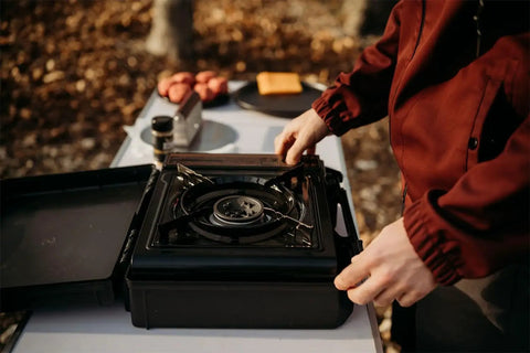 10,000 BTU Dual Fuel Camp Stove for Butane and Propane Portable Outdoor Grill on a table, person setting up for use.