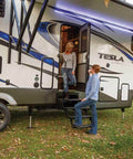 Two people by the stairs of a parked RV trailer with blue stripes, surrounded by grass and trees, engaging in conversation.
