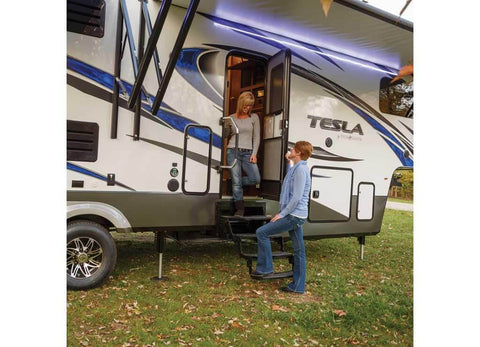 Two people by the stairs of a parked RV trailer with blue stripes, surrounded by grass and trees, engaging in conversation.