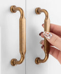Hand holding one of the ten pack antique brass cabinet pulls with three and three quarters inch hole centers, shown on a white cabinet.