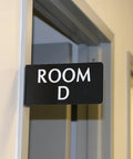 Heavy Duty Horizontal Warehouse Aisle Signs Custom Engraved L Shaped Rack Markers With Kickass Durable ABS Plastic Build Pre Drilled Mounting Holes Ready To Hang Made In Usa hanging on a door indicating Room D.