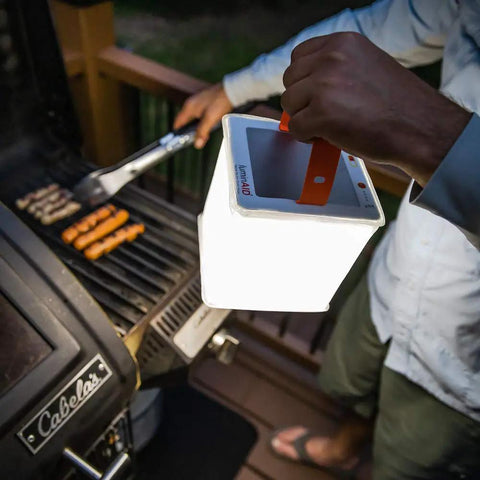 Person using 2 In 1 Solar Camping Lantern And Phone Charger With 300 Lumens Of Bright LED Light near grill at night