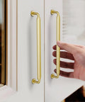 Hand touching Gold Cabinet Pulls 7.5 Inch 10 Pack Zinc Alloy Handles for kitchen cabinets with satin gold finish installed on white cabinet doors.