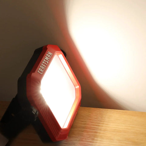 1100 Lumen LED Work Light with Tiltable Head red finish, illuminating a wall, showing impact-resistant lens and 2-in-1 handle on stand