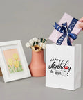 Gift scene with birthday Kraft paper bag and wrapped present, pink vase, floral art. Dope Custom Printed Kraft Paper Bags With Logo.