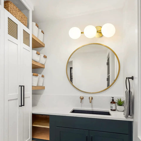 Gold Brass 3-light vanity with milk glass shades above round mirror in bathroom. Three Light Vanity Light Fixtures In Gold Brass Finish
